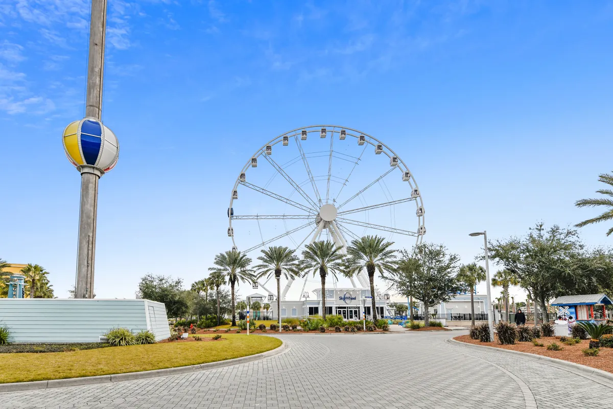 An exterior shot of a towering ferris wheel framed by palm trees, with an inviting entrance and decorative elements suggesting a festive atmosphere. The scene radiates a joyful mood, hinting at sunset rides and evening events.