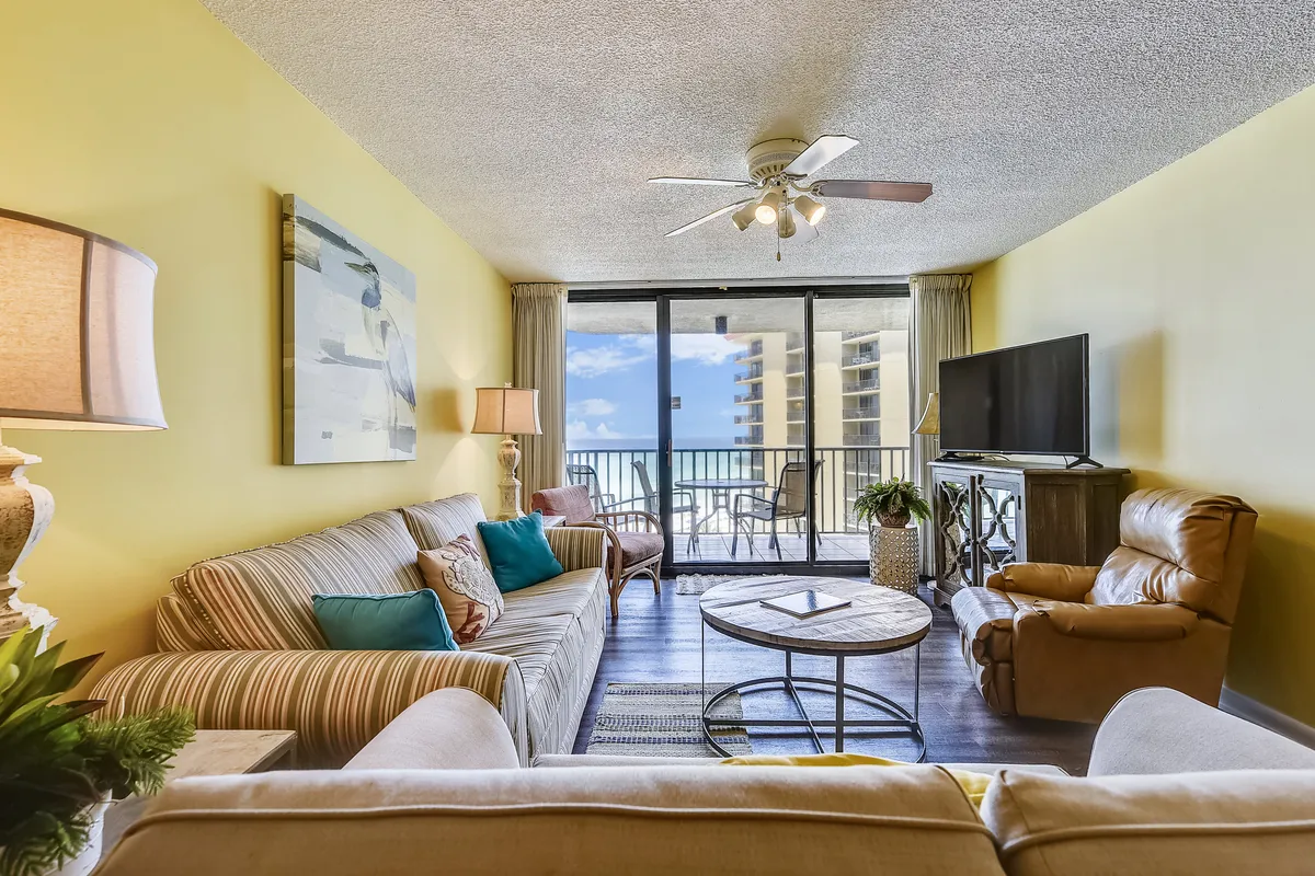 A cheerful, modern living space with a comfortable sofa, coffee table, television, and clear sightlines to the balcony. The balcony is partially visible through large glass doors, reinforcing the connection between the indoor living area and the outdoor setting.