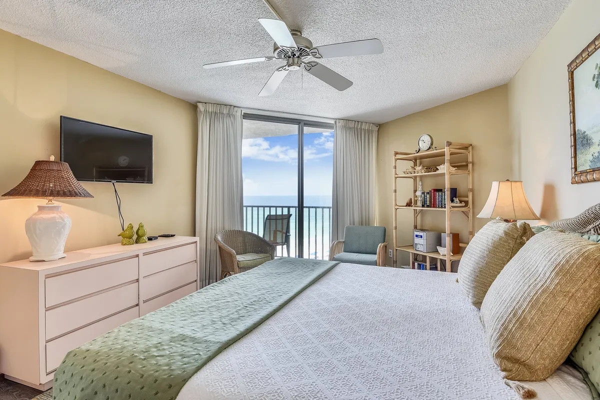A spacious master bedroom featuring a large king-size bed with plush decorative pillows, bedside tables with lamps, a wall-mounted television, a window offering an ocean view, and a bookshelf with decorative items. The soft lighting and tranquil decor create a peaceful ambiance ideal for rest.