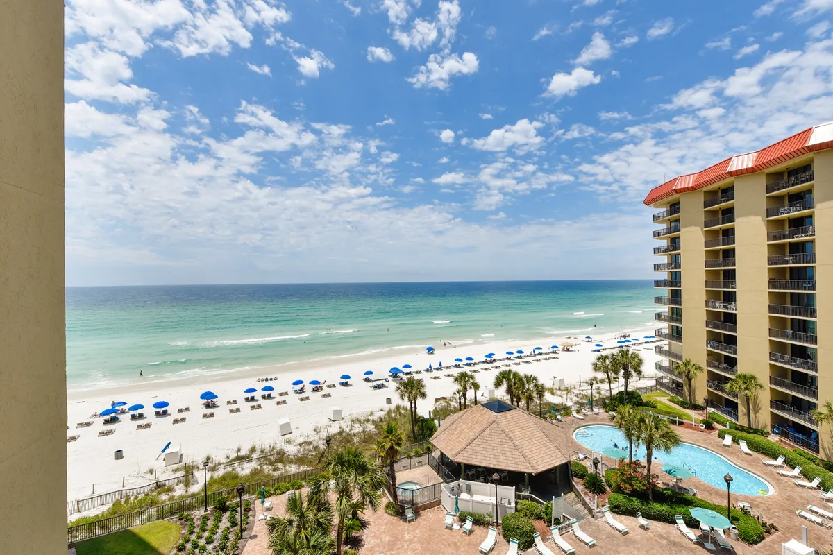 A sparkling swimming pool sits adjacent to a sandy beach lined with blue umbrellas and lounge chairs. The pool area is framed by the resort building with balconies overlooking the ocean, creating an inviting resort atmosphere with direct access to both pool and beach. Guests can be seen enjoying the water and relaxing poolside, with the panoramic ocean view in the background.