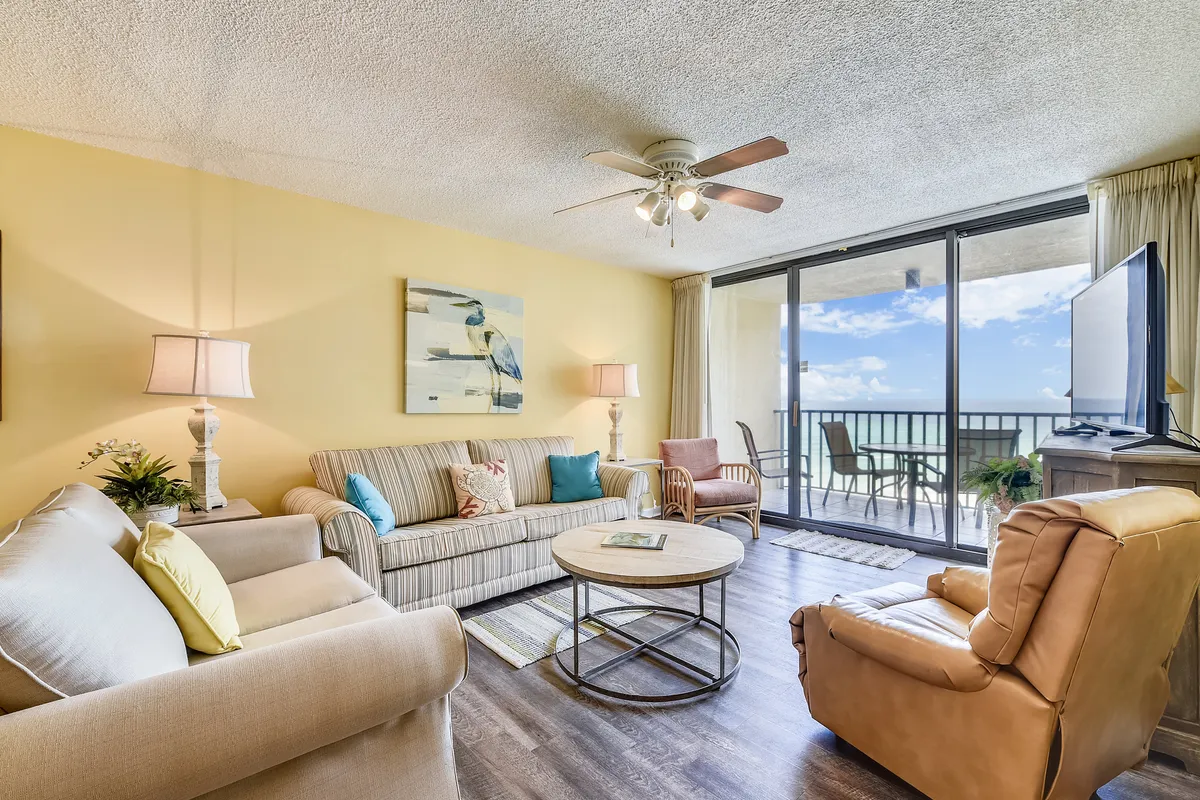 A bright and inviting living room with a comfortable sofa adorned with colorful cushions, a round coffee table, and large sliding glass doors that lead directly to a balcony overlooking the ocean. A flat screen TV sits on a wooden stand, reinforcing the space's suitability for movie nights or group gatherings.
