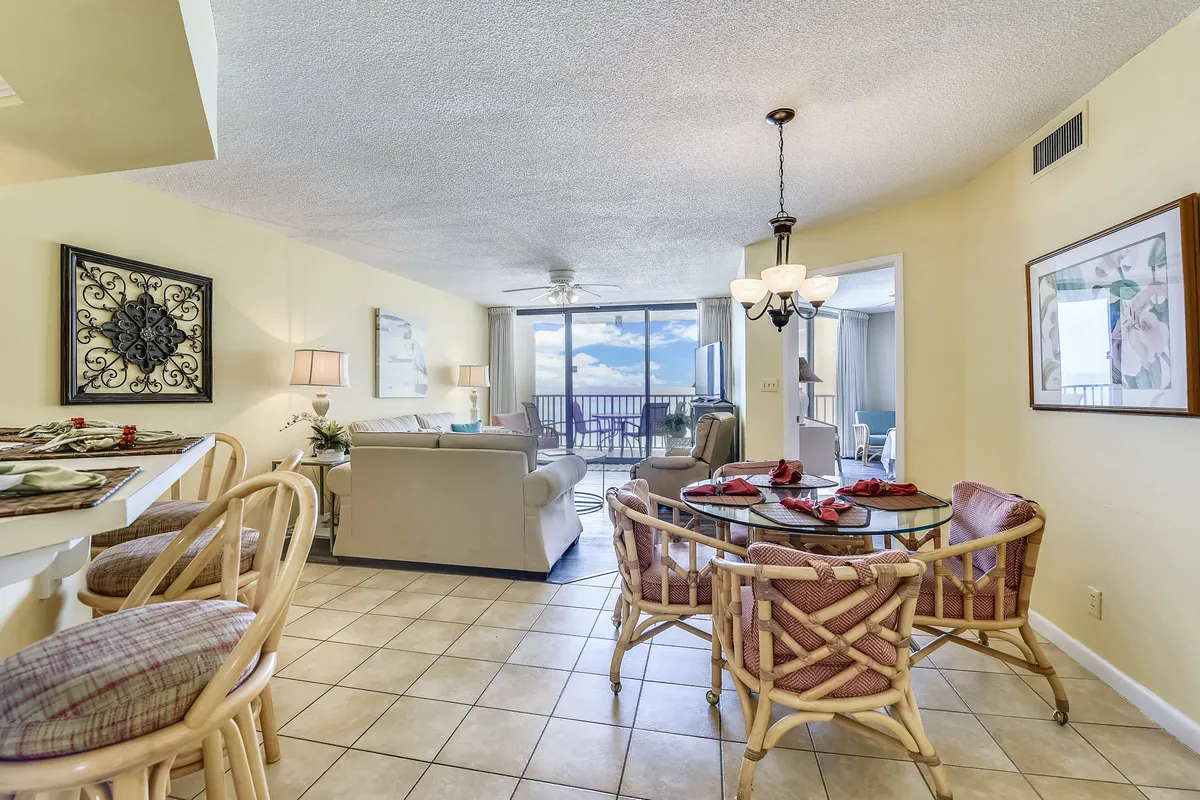 A cozy living area featuring a comfortable beige sofa facing expansive floor-to-ceiling windows, which open up to a balcony with unobstructed ocean views. The scene includes a glass dining table with cushioned wicker chairs and a kitchen counter with bar stools, all bathed in natural light. The seamless connection between the lounge and balcony is clearly visible.