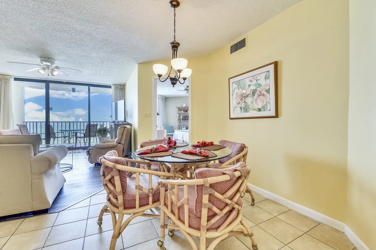 A comfortable dining area with a round table set with wicker chairs and red napkins, adjacent to a cozy living area with a sofa and armchair. Floor-to-ceiling windows reveal stunning ocean views and give access to the balcony. The entry hallway leads towards the bedrooms, suggesting an open, flowing layout.