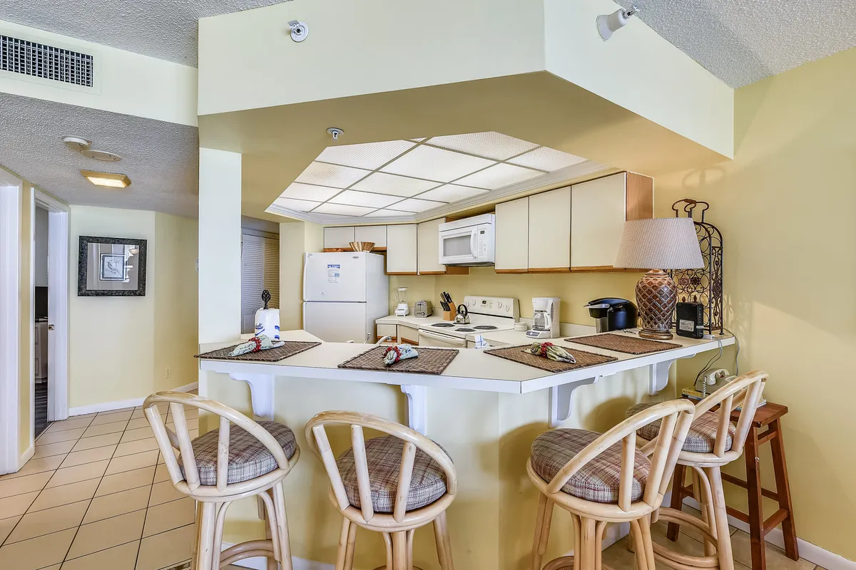 A kitchen breakfast bar with four cushioned stools, white appliances (refrigerator, stove, microwave), and a countertop set with placemats and decorative napkins. A lamp and coffee maker sit on the counter, emphasizing convenience for casual dining, breakfast, or coffee breaks. The arrangement encourages interaction and easy movement between kitchen and seating area.