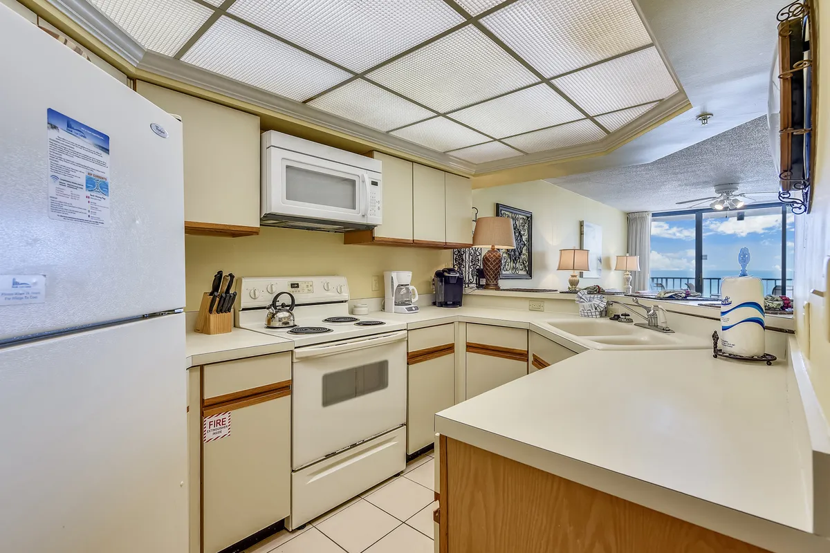 A well-lit kitchen featuring white appliances including a refrigerator, stove, and microwave. The L-shaped countertops provide ample workspace, and cabinetry offers plenty of storage. Decorative lamps and framed art add charm to the counter, while a large window with ocean view and balcony access brings in natural light and a sense of openness. The kitchen flows into adjacent spaces, suggesting an open layout ideal for socializing.