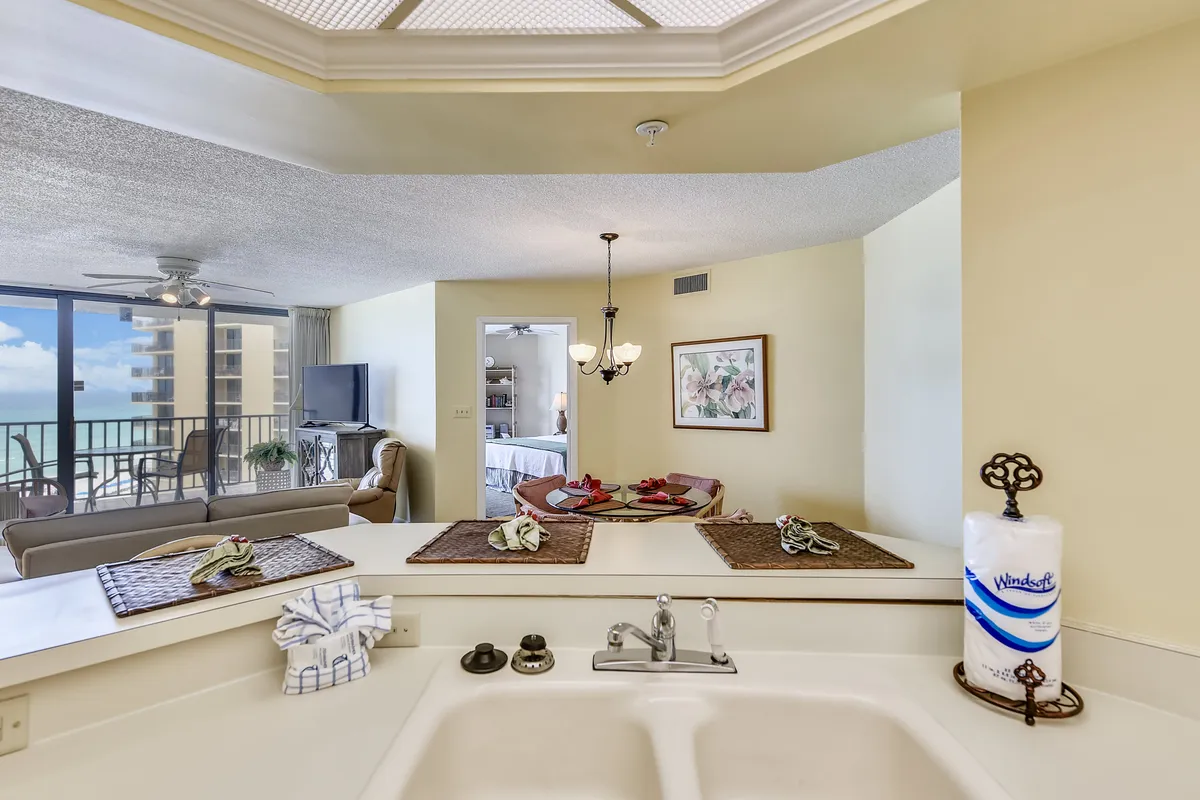 An open-plan space with a kitchen sink and counter area, dining table set with red napkins, and a living area with sofa and TV. Floor-to-ceiling balcony windows let in bright natural light and offer ocean views. The setup highlights the seamless connection between cooking, dining, and relaxing, with ample room for group activities and conversations.