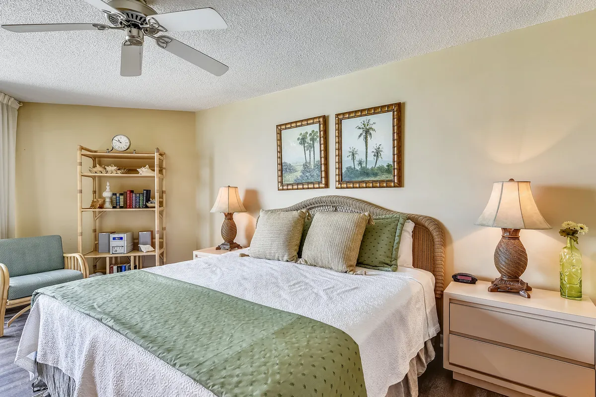 A serene master bedroom featuring a king-sized bed adorned with green and white bedding, flanked by two bedside lamps casting warm light. Bamboo-style shelving with books and decorative items adds character, while palm tree wall art reinforces the coastal theme.