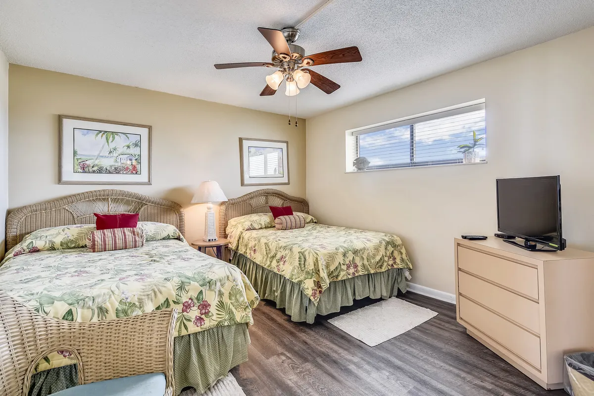 A welcoming second bedroom with two twin beds dressed in floral bedding, wicker furniture including a chair and headboards, and a ceiling fan. Natural light filters in from a small window, creating a peaceful setting ideal for kids or guests.