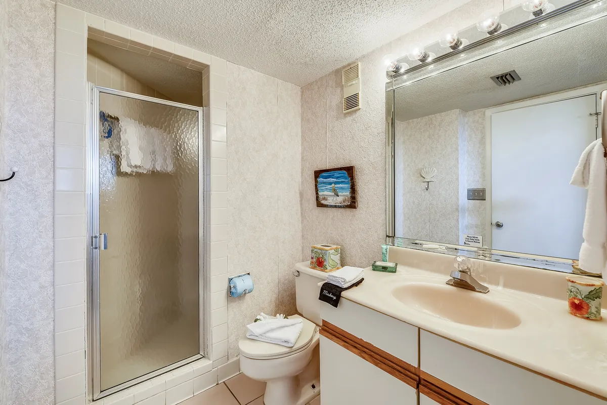 A clean, orderly bathroom with a shower featuring a textured glass door, a toilet with neatly folded towels, a large mirror with ample lighting, and a sink with floral decorative touches.