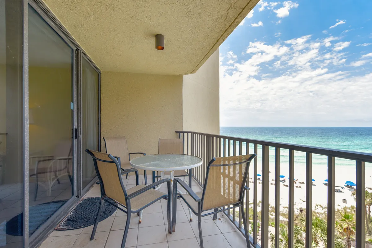 This image captures a serene seating area on a private balcony overlooking the expansive beach and ocean, with umbrellas dotting the sand and a glass sliding door connecting the balcony to the interior. The scene suggests a perfect spot for groups or families to gather, enjoy meals, or simply relax together while taking in panoramic coastal views.