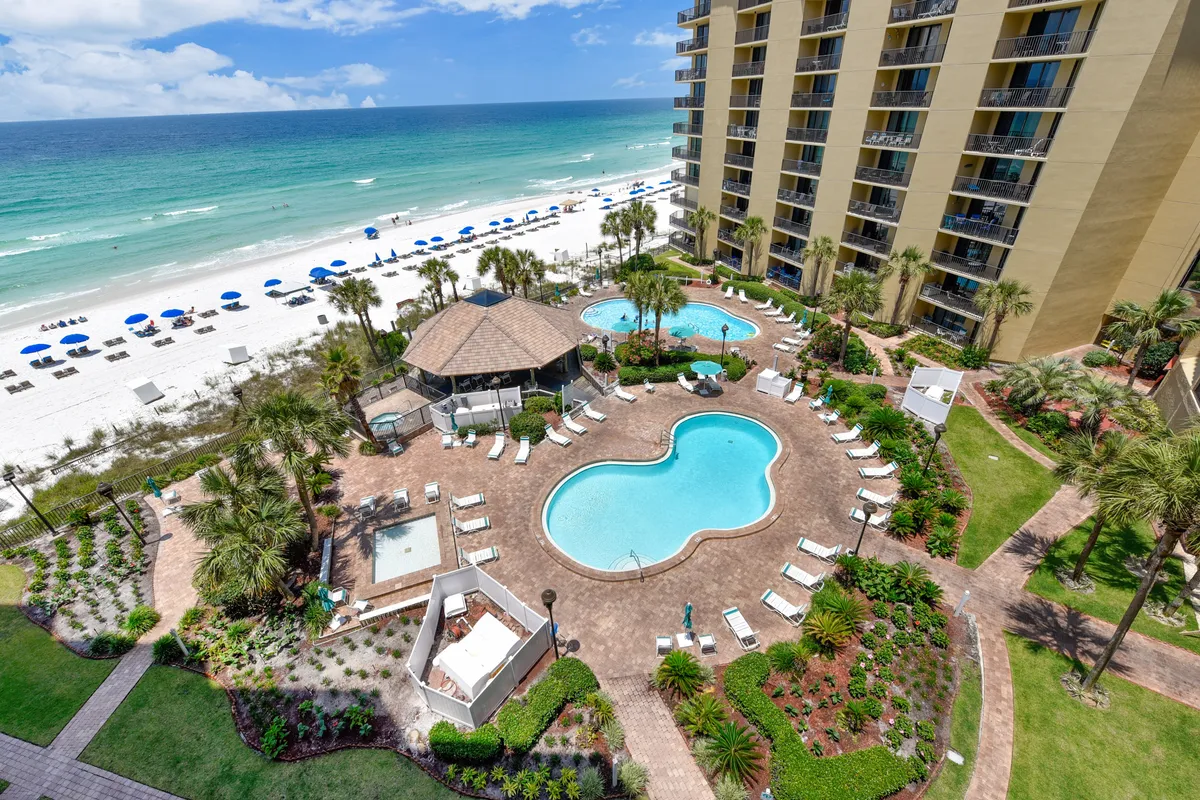 A freeform swimming pool with crystal-clear water sits beside a thatched-roof gazebo and a wide beach dotted with umbrellas and lounge chairs. The tall condominium building looms in the background, while the pool area is lively and open, ideal for social gatherings, sunbathing, and enjoying outdoor amenities.