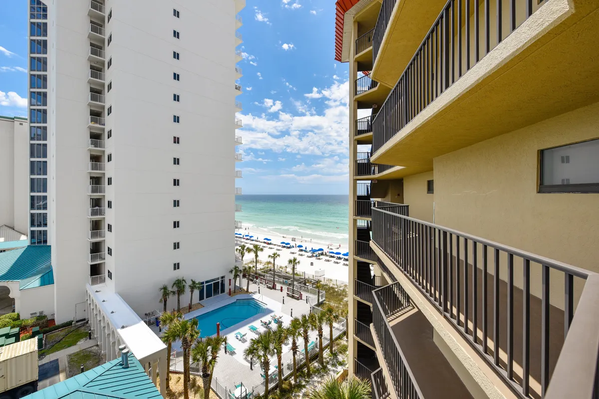 This exterior image features a sparkling community swimming pool set beside a sandy beach lined with blue umbrellas, with the panoramic ocean view stretching into the distance and balconies of adjacent buildings visible. Families can be imagined swimming, sunbathing, or strolling along the shore, all within steps of their accommodation.