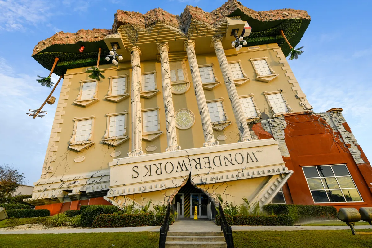 This image features the whimsical upside-down building of WonderWorks, complete with classical columns and lush green landscaping. The unique architecture immediately draws attention and invites curiosity about the hands-on science and entertainment exhibits inside.