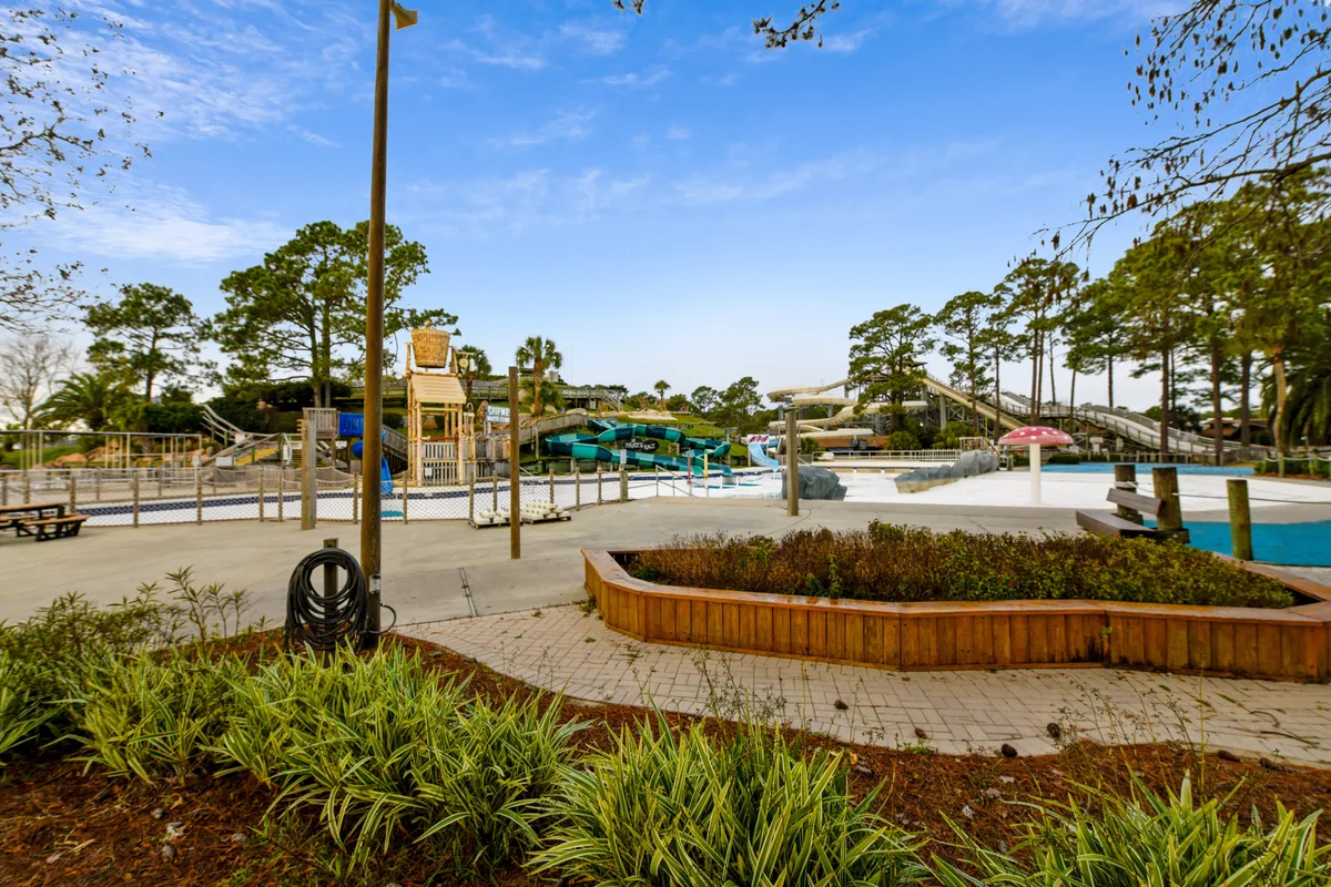 A water park scene with multiple colorful water slides, a pool area featuring a mushroom fountain, wooden planters with lush greenery, and paved walkways lined with benches. The area is vibrant and designed for fun and relaxation, with plenty of space for families to enjoy together.