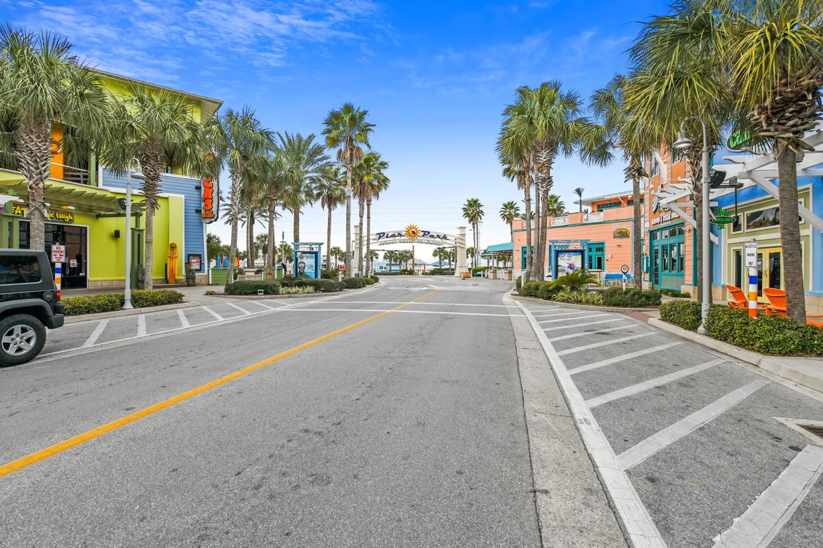 Palm trees line a cheerful street filled with colorful storefronts and outdoor dining areas, with the Pier Park archway and the towering Skywheel visible in the distance under a bright blue sky.