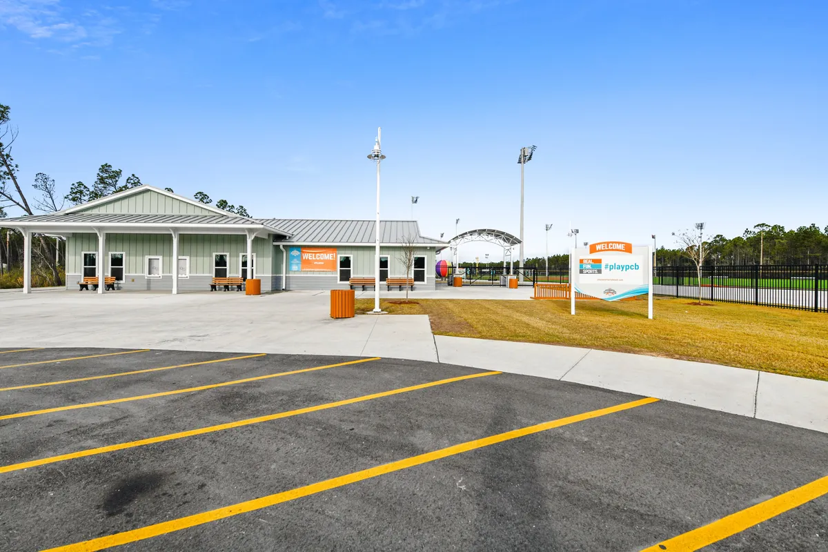 A lively outdoor scene showing bright orange 'Welcome' signs, a large pavilion, an open paved parking area, and a grassy space. The area radiates a family-friendly vibe and is set up for community gatherings, outdoor sports, and special events.