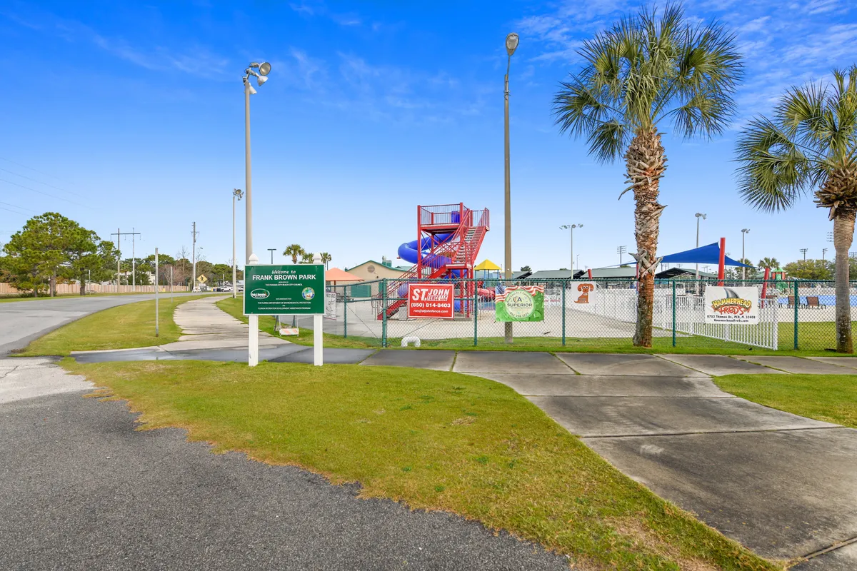 This exterior shot features a colorful water slide structure, a green entrance sign for Frank Brown Park, palm trees, and a fenced splash pad area. Children and families are implied to be enjoying the water features and park amenities, highlighting opportunities for group fun and recreation just minutes from the resort.