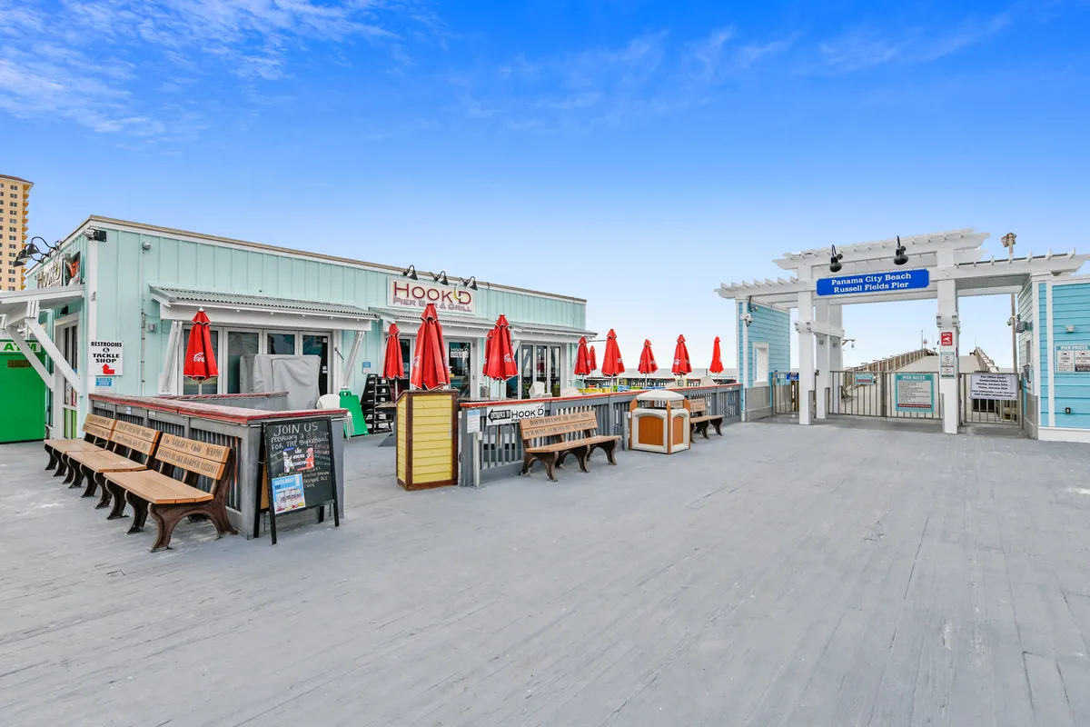 The Hook'd Pier Bar and Grill features inviting red umbrellas and open-air seating, set against the Panama City Beach Russell Fields Pier entrance. Benches line the pier, offering a relaxed, social atmosphere with ocean views.