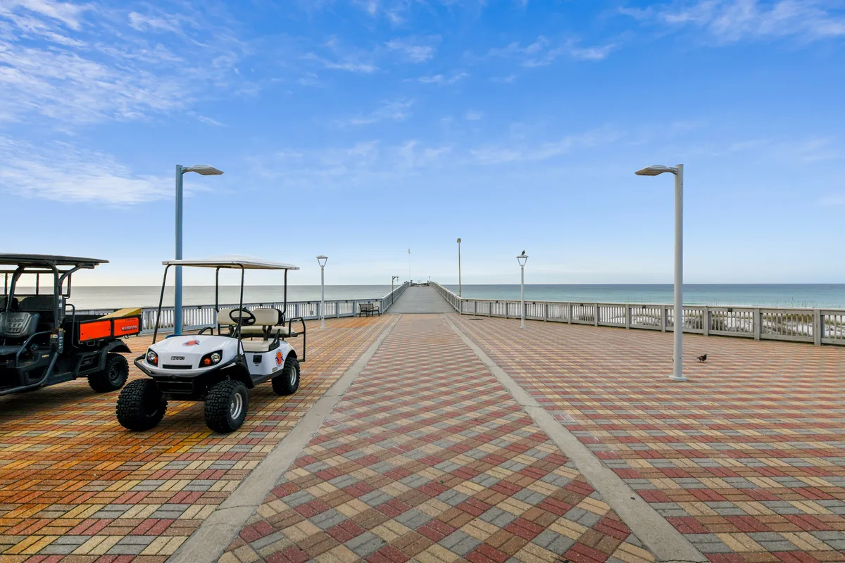 A long pier stretches into the ocean with a patterned brick walkway in the foreground, and two parked golf carts add a casual, leisurely vibe. The ocean horizon creates a sense of openness and tranquility.