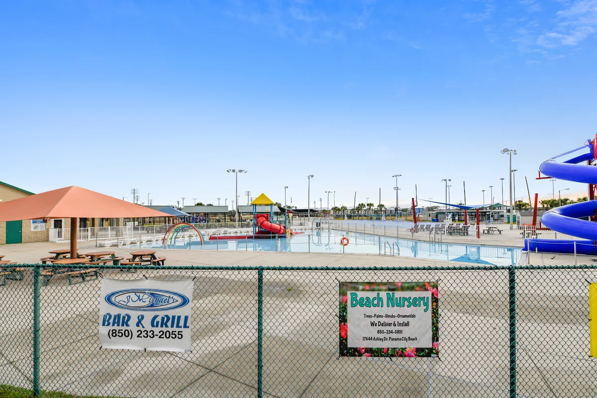 A large swimming pool with water slides is surrounded by colorful playground structures, covered picnic tables, and a fenced area, creating a playful and lively atmosphere. The area is busy with children and families enjoying both the water and the playground, making it perfect for all ages.