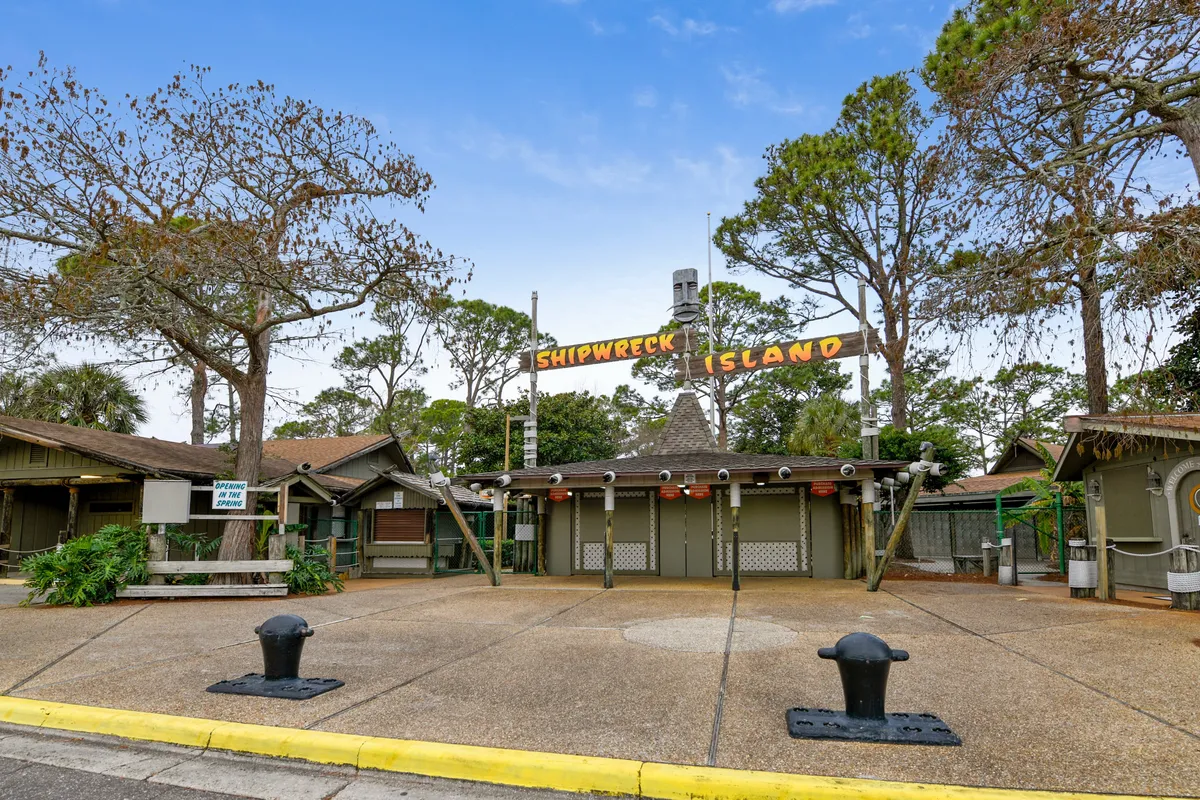 The image shows the inviting entrance sign for Shipwreck Island Water Park, surrounded by rustic wooden buildings and large trees. There’s an air of excitement and anticipation, with the themed entrance promising adventure and water park fun for families.